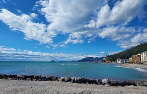 Une plage côtière bordée d’un rivage rocheux, avec un ciel bleu dégagé et des montagnes à l’horizon.