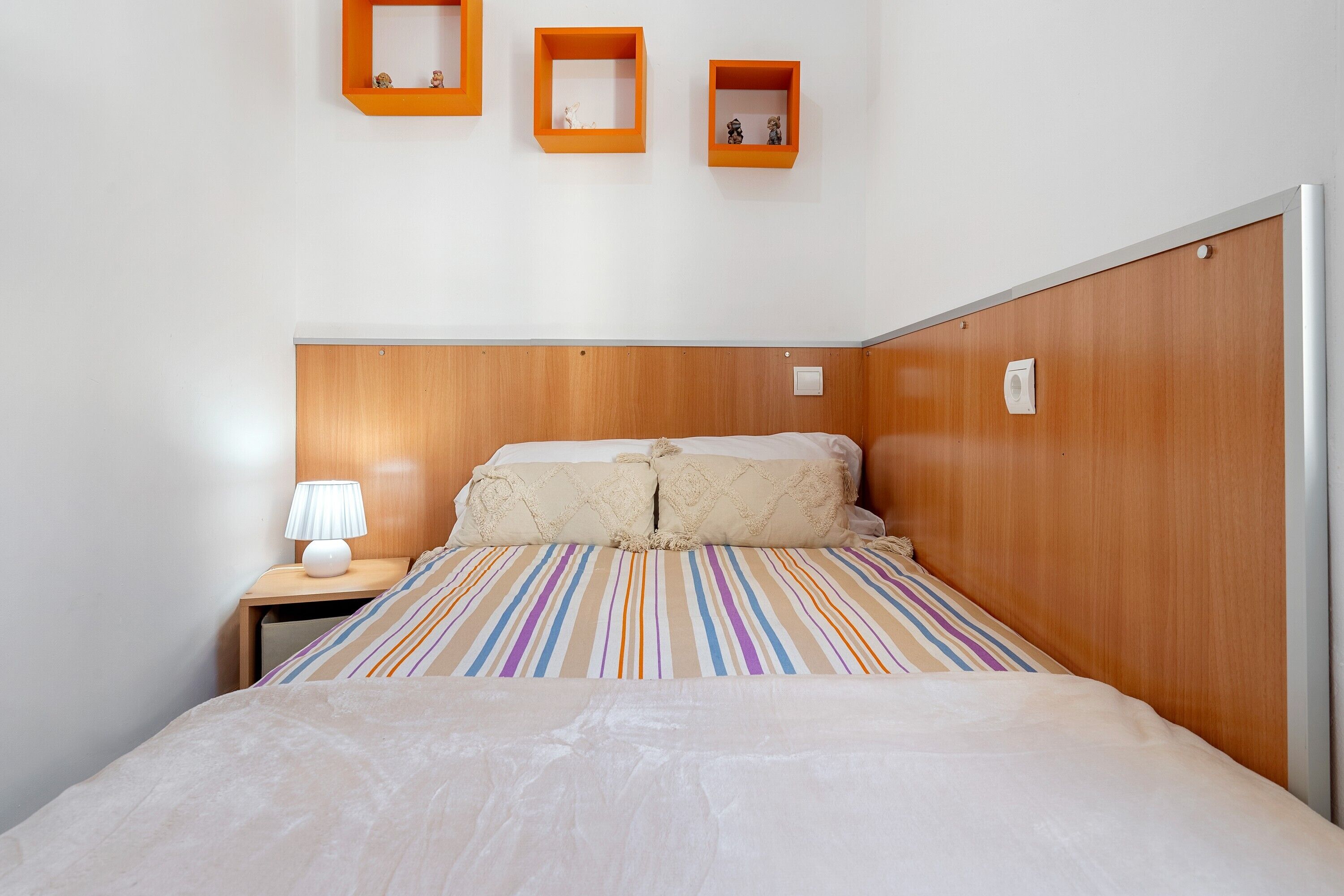 Una cama bien hecha con una colcha a rayas, una lámpara de noche y tres estantes en la pared con marco naranja.
