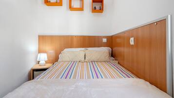 Una cama bien hecha con una colcha a rayas, una lámpara de noche y tres estantes en la pared con marco naranja.