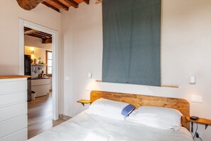 Une chambre avec une tête de lit en bois, un lit avec une literie blanche, une table de chevet, une lampe fixée au mur et une vue sur la cuisine.