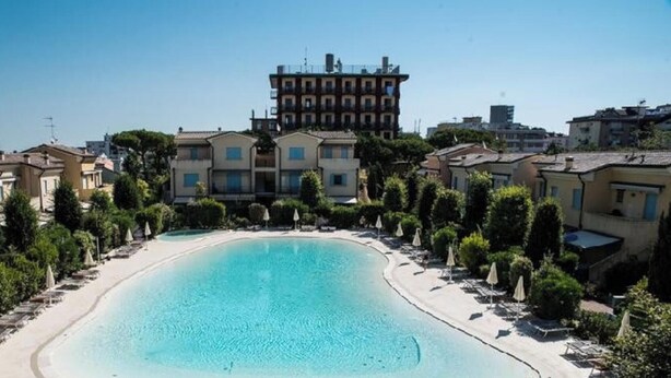Una grande piscina esterna circondata da lettini e ombrelloni, con edifici sullo sfondo.