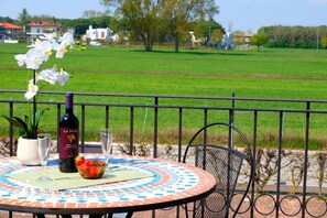 A round table with a patterned tablecloth, a bottle of wine, a bowl of fruit, and white flowers. - Apartment 'Residence Borgo Dei Fiori D03' with Shared Pool, Private Terrace and Air Conditioning (Jesolo)