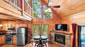 A cabin-style kitchen with wooden walls, a stone fireplace, a dining table with chairs, a large TV, and a ceiling fan.