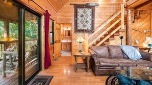A wooden cabin interior with a wooden staircase, a leather sofa, a glass coffee table, and a wooden deck with outdoor seating.