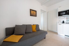 A grey sofa with a mustard throw, a framed picture on the wall, and a kitchen area with white cabinets and appliances.
