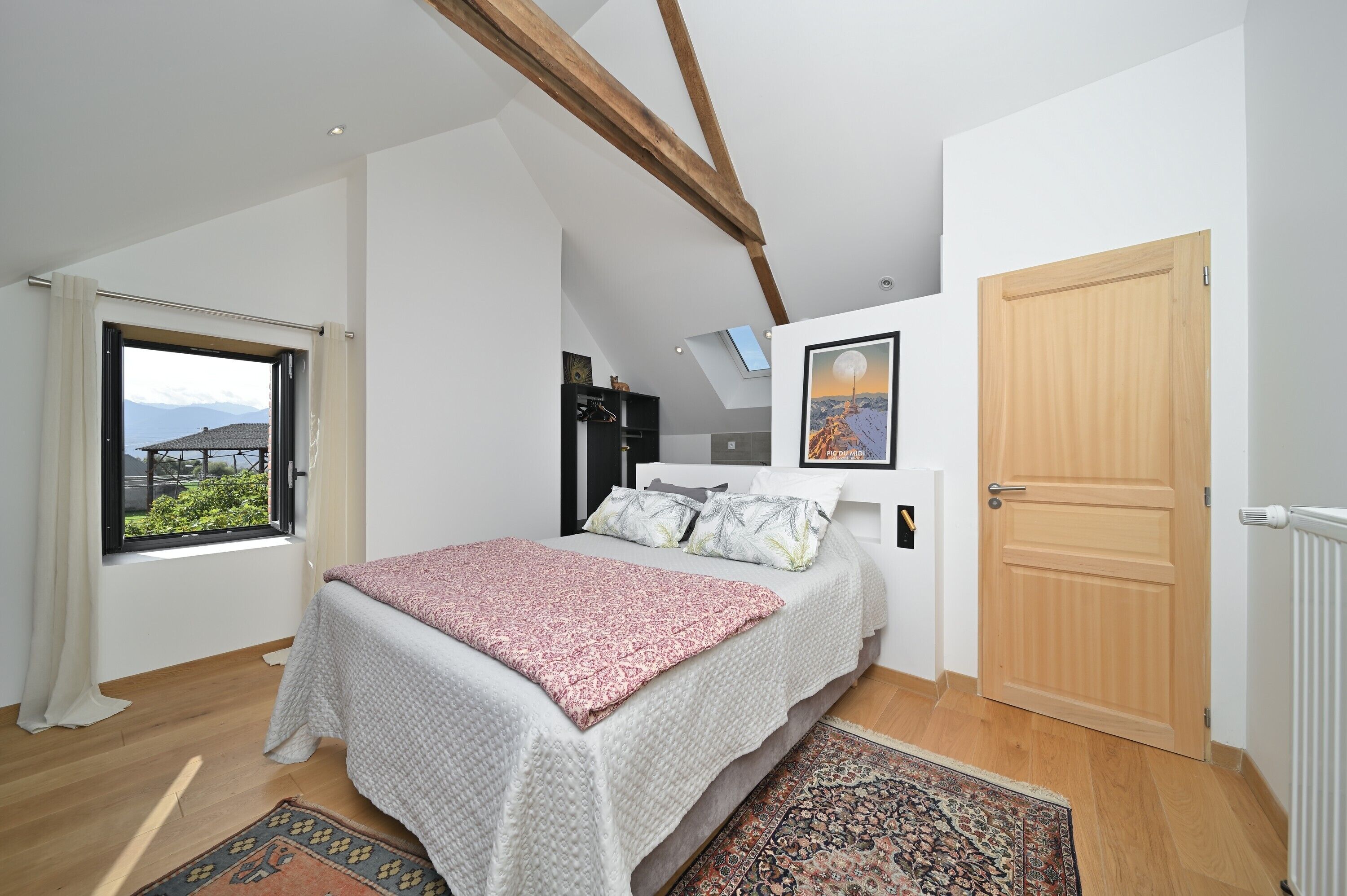 Een slaapkamer met een bed, dakraam, houten deur en uitzicht op bergen.
