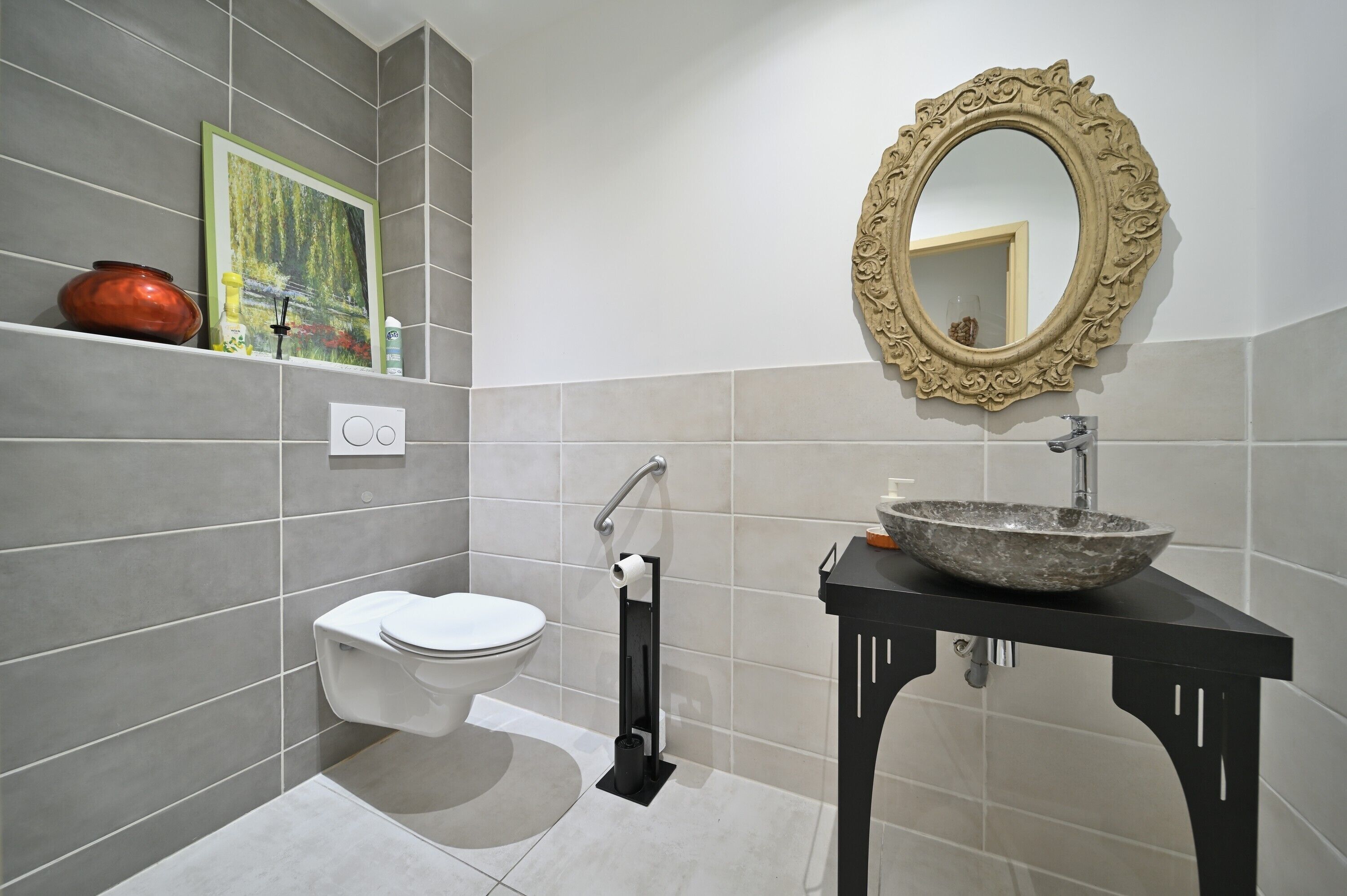 Ein Badezimmer mit Toilette, Waschbecken, Spiegel und Ablagefläche mit Vase und Bilderrahmen.