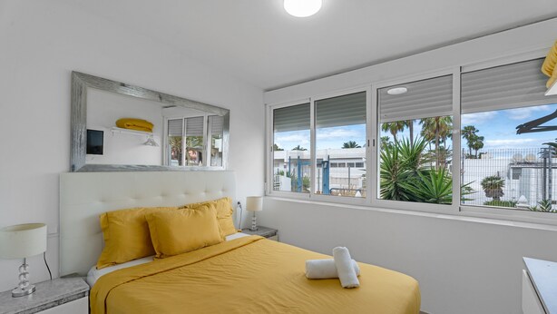 A hotel room with a bed, a nightstand, a lamp, a mirror, and a window with a view of palm trees and buildings. - Vacation Apartment 'Costa Del Silencio Tenerife Sur' with Shared Pool and Wi-Fi (Arona)