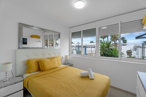Una camera d'albergo con un letto, un comodino, una lampada, uno specchio e una finestra vista palme e edifici.