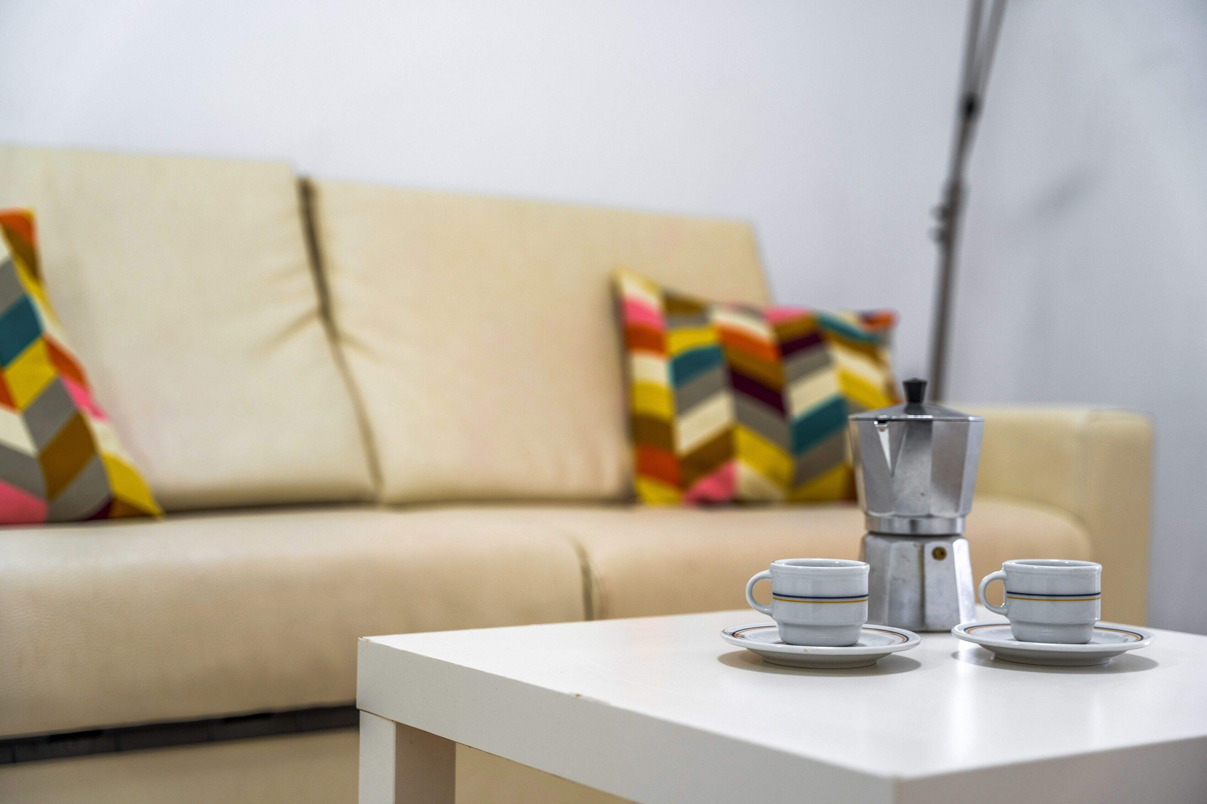 Ein weißer Tisch mit zwei Tassen und einer Kaffeemaschine in einem Wohnzimmer mit einem beigen Sofa und bunten Kissen.