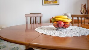 Una mesa redonda de madera con un posavasos de encaje, un frutero con plátanos, manzanas y naranjas, una silla y un jarrón en la pared.