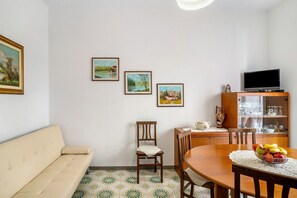 Una sala de estar con sofá, mesa de comedor de madera, sillas, televisor en un mueble y cuadros en la pared.