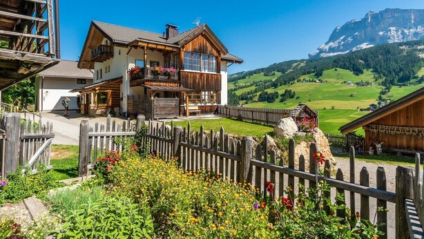 Un bâtiment de style chalet doté d’un balcon, entouré d’une clôture en bois et d’un jardin où poussent différentes variétés de fleurs.
