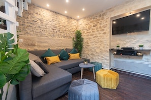 A modern living room with a stone wall, a grey sectional sofa, a small round table, a yellow ottoman, and a flat-screen TV mounted on the wall. - Apartment 'Duplex Parisien Charmant Métro13' with Wi-Fi (Paris)
