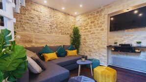 A cozy living room with a stone wall, a grey sofa, a small round table, and a yellow ottoman. - Apartment 'Duplex Parisien Charmant Métro13' with Wi-Fi (Paris)