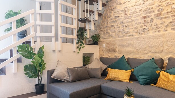 A modern living room with a grey sofa, yellow ottoman, and a staircase with white railings. - Apartment 'Duplex Parisien Charmant Métro13' with Wi-Fi (Paris)