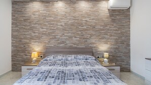 A bedroom with a patterned bedspread, two bedside lamps, a wall-mounted air conditioner, and a wooden headboard. - Villa with Jacuzzi & Fitness Area – Private Spaces & Exclusive Areas – Arenella Mare (Arenella)