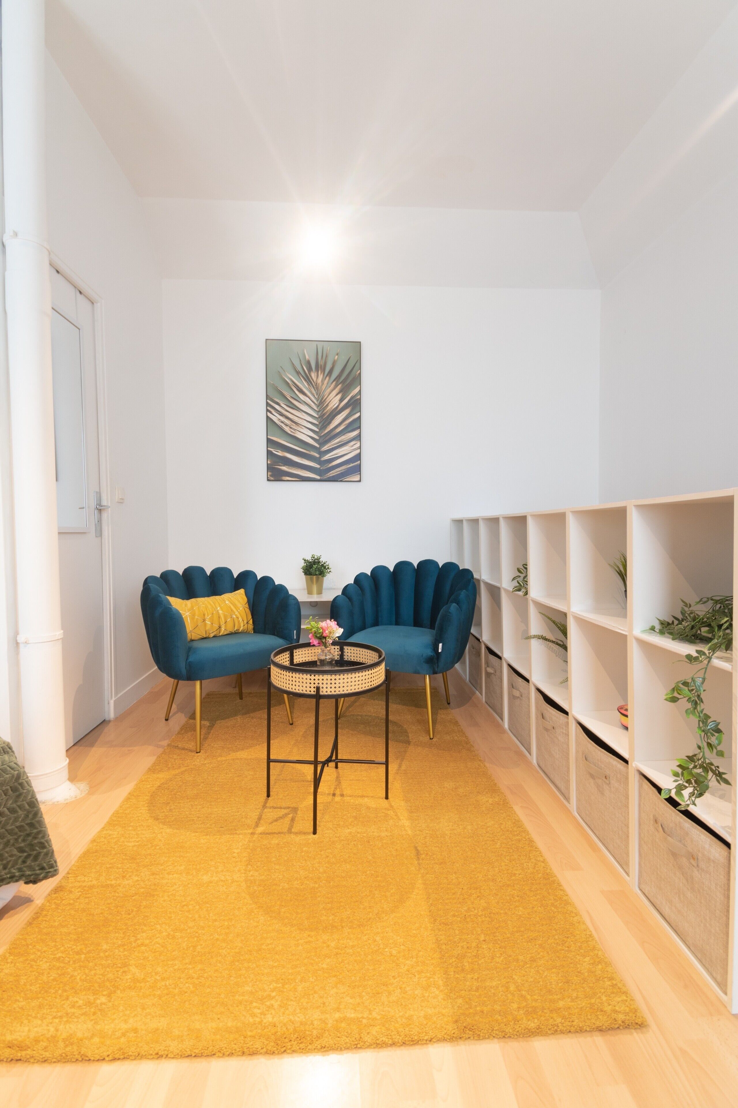 A room with a yellow rug, two blue armchairs, a round coffee table, and a white shelving unit with plants.