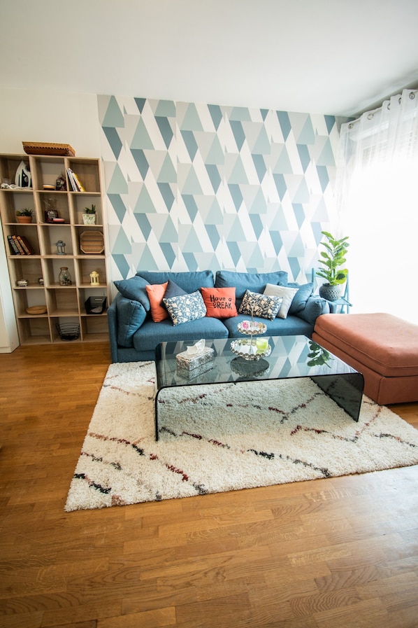 A living room with a blue sofa, a glass coffee table, a patterned rug, and a geometric wall design.