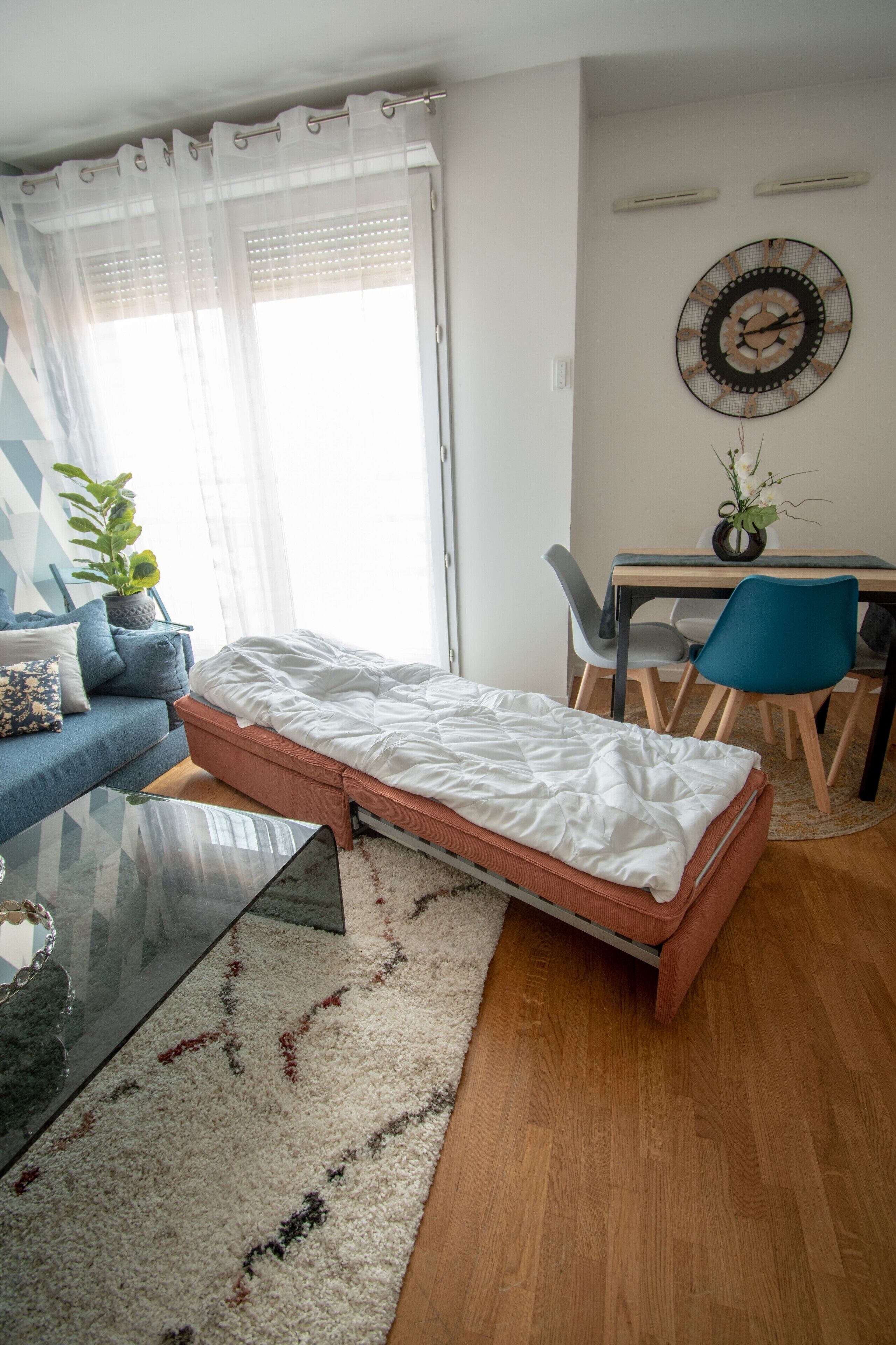 A compact living space with a bed, a sofa, a glass coffee table, a desk with chairs, and a wall clock.