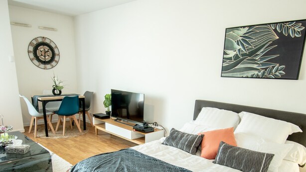 A bedroom with a bed, bedside table, TV, desk with chair, and a wall clock.
