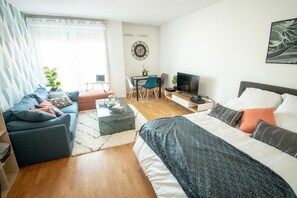 A modern, well-lit room with a sofa, a bed, a dining table with chairs, a TV, and a small desk.
