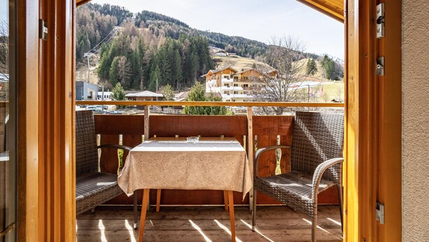 Un balcón de madera con mobiliario de mimbre y una mesa, con vistas a un paisaje montañoso con edificios.