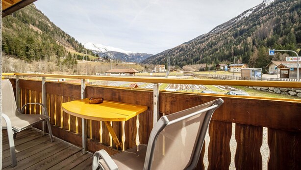 Une terrasse en bois avec une table et des chaises jaunes, offrant une vue sur un paysage montagneux avec une voie ferrée et des bâtiments.