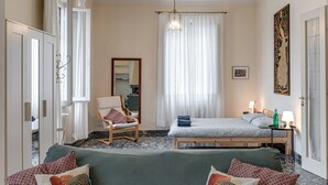Makuuhuoneessa on sänky, tuoli, sohva, peili, ikkuna, jossa on verhot, yöpöytä lampun kanssa ja taulu seinällä.