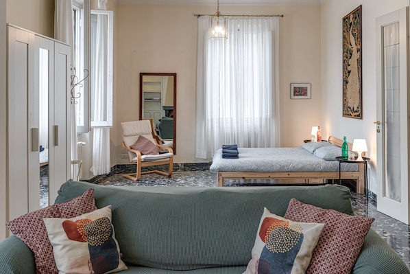 A bedroom with a bed, a chair, a sofa, a mirror, a window with curtains, a bedside table with a lamp, and a painting on the wall.