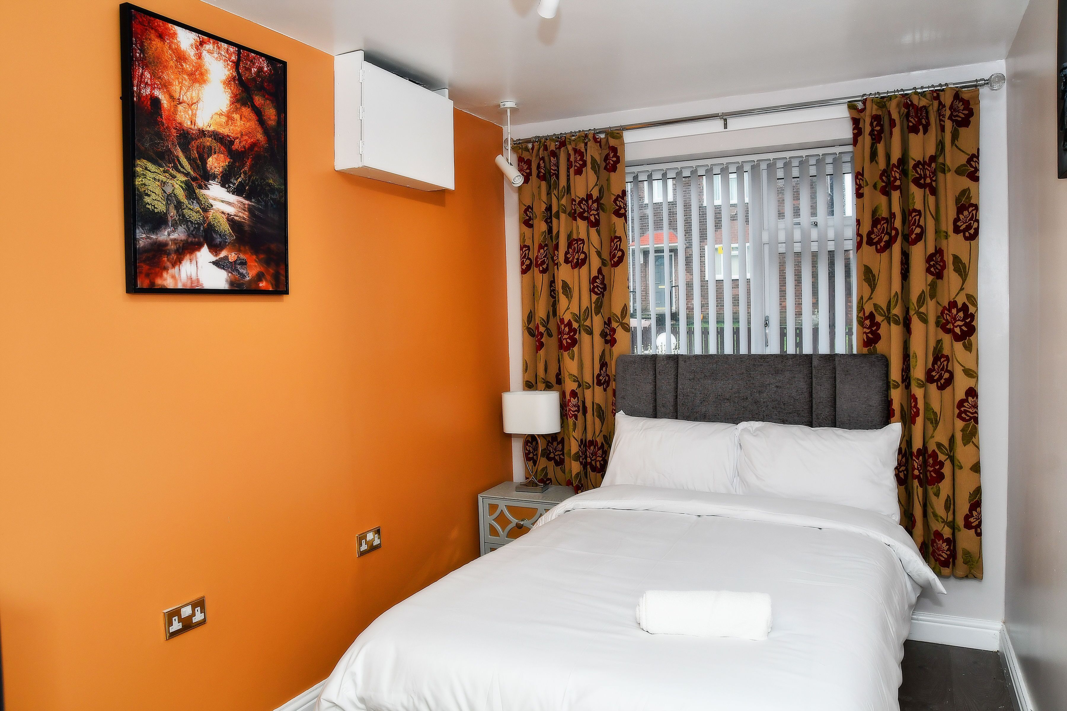 Une chambre d’hôtel avec un mur orange, un lit avec une literie blanche, une tête de lit grise et un rideau à motifs floraux.
