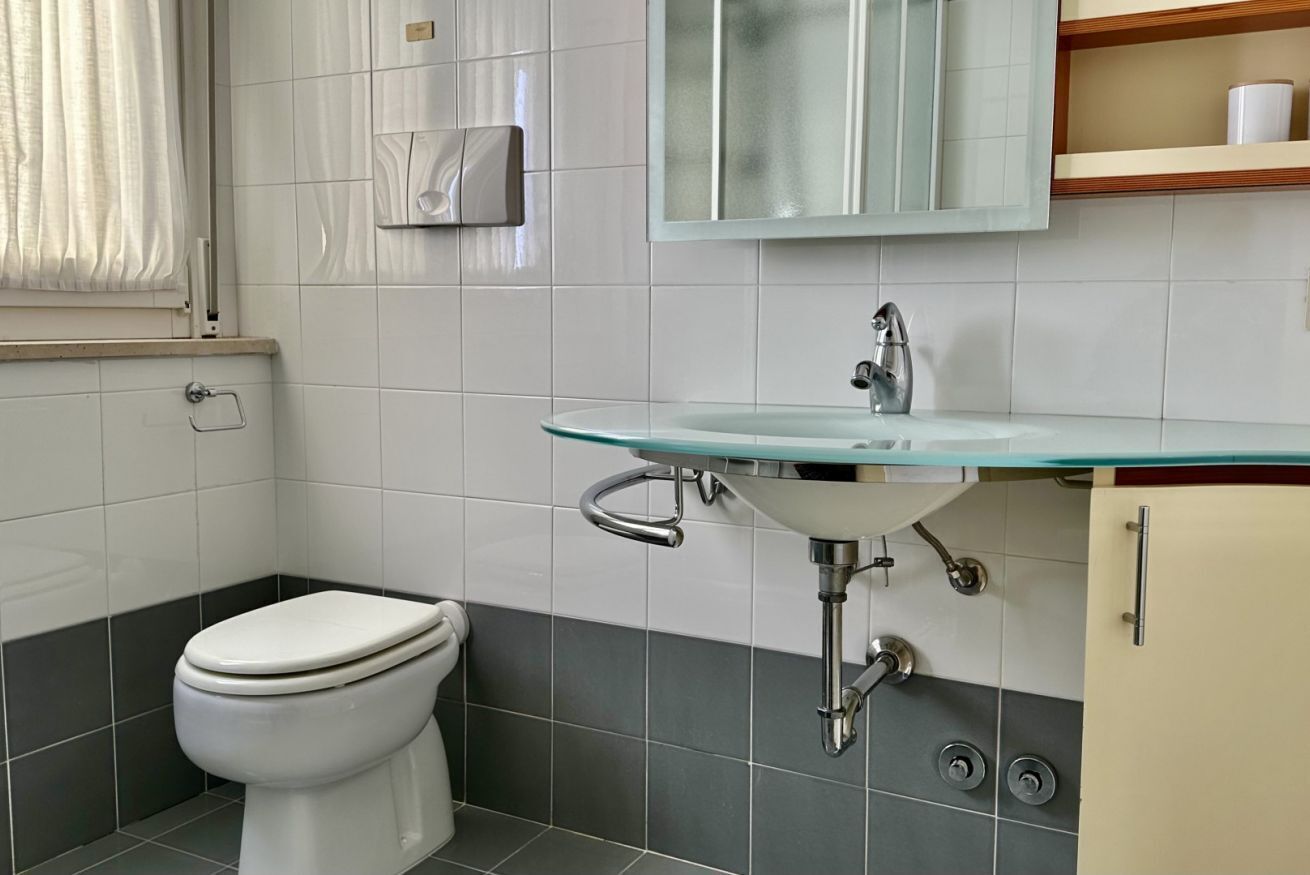 A bathroom with a toilet, a glass sink, a mirror, and a towel dispenser.