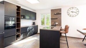 A modern kitchen with dark cabinetry, a built-in oven, a coffee machine, and a large window.