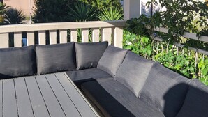 A sectional sofa with a wooden table, a white railing, and greenery in the background.