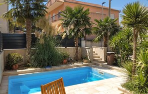 A swimming pool with a wooden chair, surrounded by various plants and a building with balconies. - Awesome home in Podstrana (Podstrana)