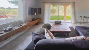 A modern living room with a large window, a flat-screen TV, a wooden bench, and a dining area with a table and chairs. - Luxurious home in Mouries with private swimming pool, can be inside or outside (Mouries)