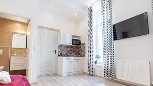 Quarto de hotel moderno com cozinha compacta, cama, televisão e janela com cortinas estampadas.