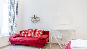 Quarto com sofá vermelho, escrivaninha branca, prateleira pequena com plantas e janela com cortinas estampadas.
