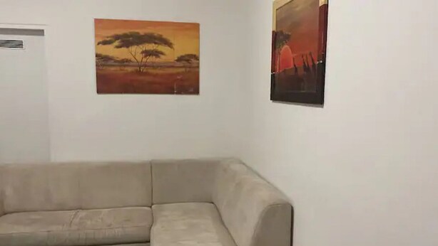 A beige sectional sofa in a room with white walls, two paintings depicting landscapes, and a vent on the left wall. - Cozy 3BR Apt. Wien Simmering (Wien)