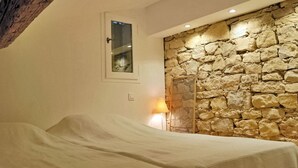 Un dormitorio con dos camas, una pared de piedra, un ventanal pequeño y una lámpara.