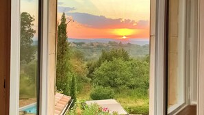 Vista a través de una ventana de un atardecer sobre un paisaje montañoso con vegetación y una piscina visible en primer plano.