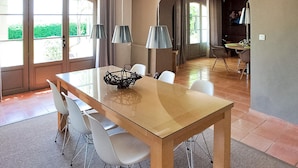 A dining area with a wooden table, white chairs, and a glass-top table. There are two lamps on the table and a basket on the left side. The room has large windows with curtains, a tiled floor, and a view of greenery outside. - Amazing home in Violes (Violes)