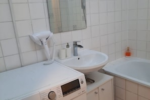 Un baño con un lavabo blanco, un espejo, un secador de pelo de mano y un reloj digital.