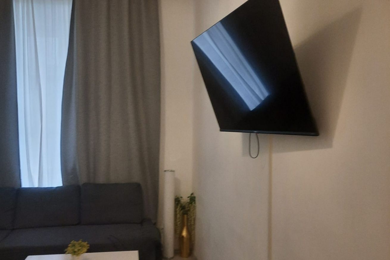 Ein Wohnzimmer mit Flachbildfernseher an der Wand, einer Couch und einem kleinen Tisch mit einer Vase.