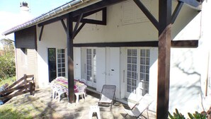 Een overdekt terras met een tafel en stoelen, een houten hek en een wit gebouw met ramen en deuren.