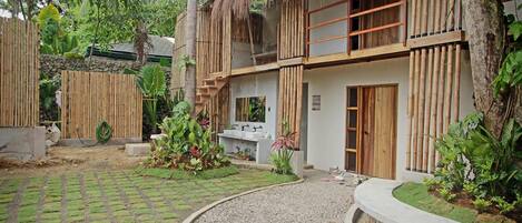 A two-story building with a thatched roof, surrounded by lush greenery and a paved courtyard.