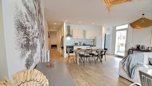 A modern living room with a dining area, a kitchen in the background, and a large window providing natural light. - Pen-guen - Saint Cast LE Guildo (SAINT CAST LE GUILDO)