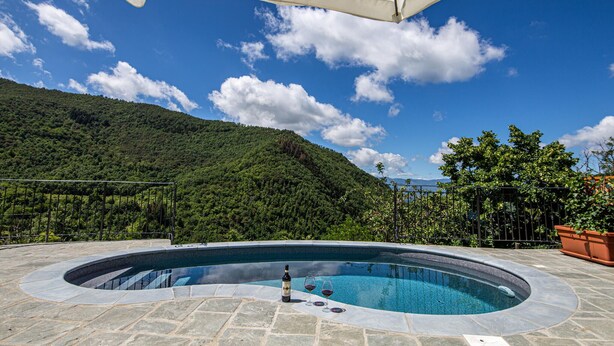 Una piscina con borde de piedra, rodeada por una zona de patio con una sombrilla blanca, con un exuberante paisaje de colinas verdes de fondo bajo un cielo azul despejado.