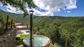 Un jacuzzi con revestimiento de madera, rodeado por un muro de piedra y una terraza de madera, con vistas a un valle verde y exuberante con montañas al fondo.
