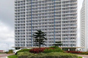 A high-rise building with a uniform facade, multiple balconies, and a landscaped area with shrubs and a small tree.